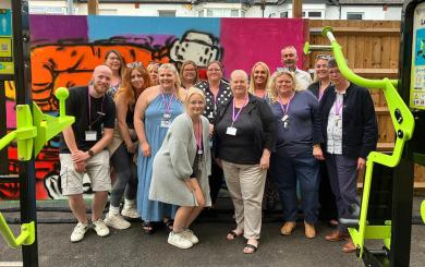 A group of male and female Sanctuary Supported Living staff stand in front of a graffiti mural, smiling. Most wear purple lanyards, and lime green gym equipment like a Lat Press is visible in the foreground.