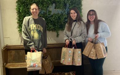 Three white women in their 30s standing inside the entrance to a pale-walled modern church, with a 'River Church' sign behind them. They're smiling at the camera and surrounded by brown paper Christmas gift bags laid out across the floor.