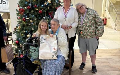 An older lady wearing a blue dress and white cardigan seated by a large Christmas tree in the bright atrium of a retirement community, with a young blond-haired girl next to her, holding a large Christmas gift bag. Behind them is a lady in her 30s wearing a white blouse and purple lanyard, with a lady in her late 50s or 60s stood next to her. All are smiling at the camera.