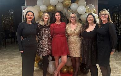 Six white women aged from their 20s to their 50s, all dressed in black, red or gold party dresses, stood inside a retirement community large communal room, on top of a red carpet leading to a white and gold balloon arch . They're stood together smiling at the camera.