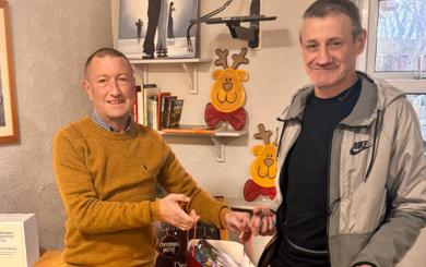 Two men standing together, the first wearing a mustard-colored jumper and dark wash jeans, and the second, taller man in a Nike tracksuit top. They are holding Christmas gift bags and smiling.