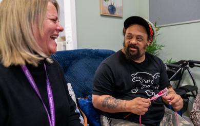 A white woman with blonde hair wearing a purple Sanctuary Supported Living lanyard laughs next to a man with darker skin, wearing a rastacap, sideburns, and a beard, using crochet needles with pink and white wool