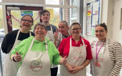 A group of six people standing together indoors, all wearing white aprons with the "Fresh Beginnings" logo. They are smiling and posing for the camera. One person is giving a thumbs-up, while another is making a peace sign. They appear to be in a kitchen or community space.