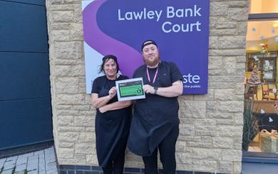 Two members of a kitchen team standing outside Lawley Bank Court, as shown by a purple sign on the building, holding a certificate displaying a '5' food hygiene rating. They are wearing black uniforms and smiling.
