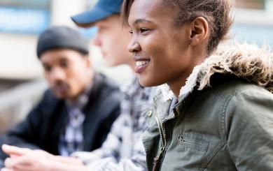 A young, black woman in a green jacket with a fur-lined hood sits outdoors smiling, while two out-of-focus young, white men in casual clothing talk in the background.