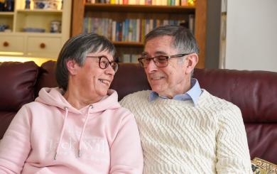 Two people wearing cosy jumpers looking into each other's eyes while sitting on a dar red sofa