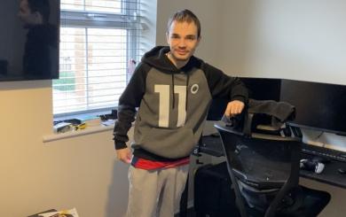 A young man with light skin, short brown hair, and a slight smile stands in a lounge area, leaning on a black gaming chair