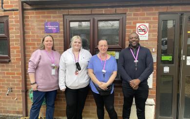 Three white women and one black man standing in a row outside by the front door of a residential care home. They are all wearing purple lanyards and are smiling at the camera. There is a green sticker in the window behind them, showing a rating of five from the Food Standards Agency.