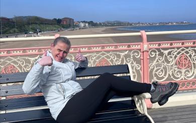 A woman wearing a pale grey jacket, black leggings and trainings, sits along a bench on a pier by a beach, smiling and giving a thumbs up to the camera.