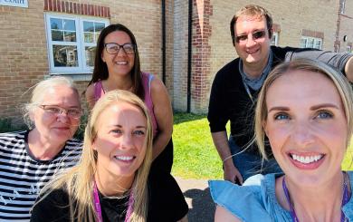 Four women and one man, wearing purple Sanctuary Supported Living lanyards are standing on a lawn outside a large brick building, posing together and smiling for the camera.