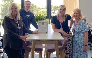 Three women and one man stood around an office table, smiling at the camera and showing off a certificate in the middle of the group