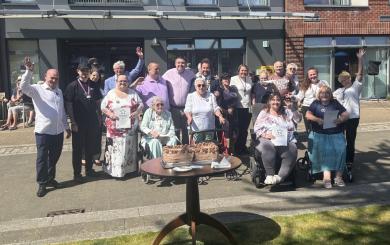 A large group of people stood or sat in wheelchairs all together, posing for the camera. They're in front of the main entrance to a large building, with a table in front of them which has a large rectangular chocolate cake on it. Some people are holding certificates.