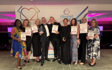 A group of nine people in suits and evening dresses, stood in a row smiling at the camera in a large room, in front of a 'Housing with Care Awards sign'. They are happily showing off their certificates and one glass trophy award.