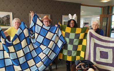 Four older people stood in a communal lounge of a retirement community, smiling at the camera and proudly displaying three knitted and crocheted blankets - one blue and cream, one yellow and green, and one purple and cream.
