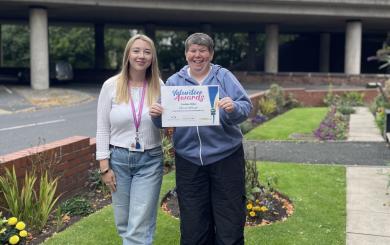 two women standing together, smiling and looking at the camera. A blonde woman on the left is wearing a white top and a purple lanyard, the woman on the right is holding up a certificate that reads volunteer awards.