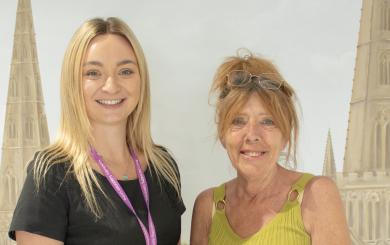 A young woman in a black dress with a lanyard stands next to an older woman wearing a green top and patterned skirt. The older woman is holding a bouquet of yellow roses. They are posing in front of a backdrop featuring tall, pointed church spires and a green landscape. Both women are smiling.