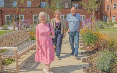 Three people walk in a sunny garden, on the left a happy older woman with glasses and short white hair wearing a pink floral dress. In the centre, a woman working for Sanctuary with brown hair, wearing a casual outfit. On the right, a cheerful man wearing a casual grey shirt and jeans. The garden is filled with colourful flowers and greenery, creating a vibrant atmosphere.