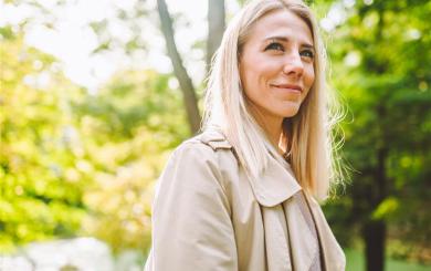 A representative photo of Lauren showing a person with long, straight blonde hair wearing a light beige trench coat, standing outdoors in a green, leafy park setting. Sunlight filters through the trees in the background, creating a bright and natural atmosphere.