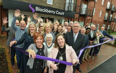 A group of people gathered outside a modern brick building with balconies, standing in front of a sign that reads “Blackberry Court.” They are participating in a ribbon-cutting ceremony, holding a long purple ribbon stretched across the entrance. Many individuals have their hands raised in celebration, and the setting appears to be the opening of a new facility or building.