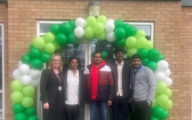 A group of five people stands in front of a Budgens store, which features a large arch made of green and white balloons. The individuals are smiling and dressed in casual clothing. The store's sign is prominently displayed above the entrance. The setting appears to be a festive occasion, likely a store opening or celebration.