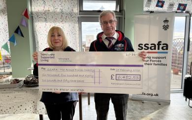 A man and a woman stand together in a bright, decorated room, holding a large novelty cheque made out to SSAFA - The Armed Forces Charity.