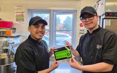 Two chefs in black uniforms and caps are standing in a kitchen. They are smiling and holding up a green food hygiene rating certificate that displays a score of 5. The kitchen is equipped with various cooking appliances and utensils, and there is a window in the background showing a view of parked cars outside.