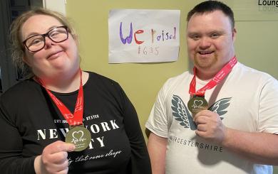 A woman and a man stand side by side, both smiling and holding medals. They are wearing red lanyards around their necks. Behind them is a sign that reads "We raised 1635," indicating a fundraising achievement.