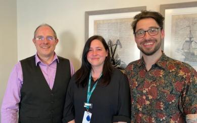 Three members of the Sanctuary Supported Living Learning and Development team standing indoors. One person wears a lilac shirt with a dark waistcoat, one wears a dark long‑sleeved top with a green lanyard, and one wears a patterned short‑sleeved shirt. They are standing side by side in front of framed artwork on a light‑coloured wall.