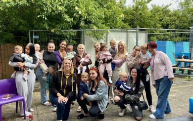 A group of women and children are gathered outdoors on a paved area. Some women are holding babies, while others are standing or kneeling with toddlers. The group is smiling and appears to be enjoying a casual gathering. In the background, there are trees, a fence, and some outdoor furniture. The atmosphere is relaxed and friendly.
