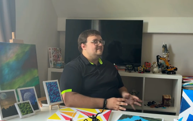 Person sitting at a desk with paintings and framed artwork, speaking in a home studio setting.