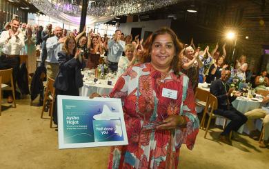 A woman in a red and white patterned dress stands smiling, holding a framed certificate. The certificate reads "Aysha Hajat, Employee of the Year." Behind her, a large group of people are standing and clapping in a decorated venue with draped fabric and lights. Tables are set with flowers and glasses, suggesting a celebratory event.