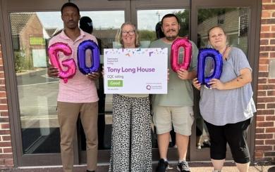 Four individuals stand in front of a building, holding balloons that spell "GOOD" and a sign with a "Good" CQC rating for Tony Long House.