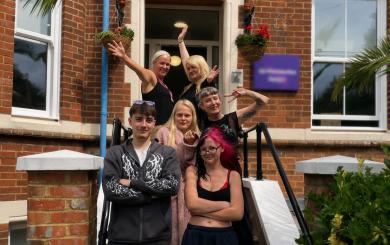 Group of six people standing on the steps outside a red brick residential building. Two people stand at the front with arms folded, while four others pose behind on the staircase. Hanging flower baskets frame the entrance with surrounding white-framed windows, black handrails and greenery.