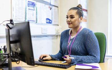 A young woman with a bun hairstyle sits at a desk, working on a computer. She is wearing a blue sweater and has a lanyard around her neck. The desk is organized with a computer monitor, a keyboard, a notepad, and a pen. Behind her, there are various documents and notices pinned to a board. The setting appears to be an office or study environment.