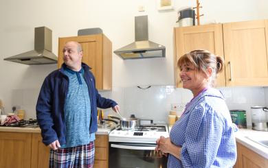 A man in a blue jacket and plaid pajama pants stands at a stove, stirring a pot. He looks to the side with a slight smile. A woman in a blue checkered shirt stands nearby, smiling back at him. The kitchen has wooden cabinets and modern appliances, with various kitchen items visible on the counter.