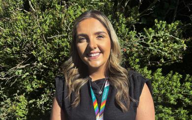 A young woman with long, wavy hair stands smiling in front of a lush green background. She is wearing a black top and a colorful lanyard with a rainbow design around her neck. The sunlight highlights her features, creating a warm and inviting atmosphere.
