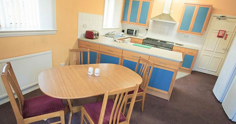 Compact kitchen with wooden units and a pop of royal blue, including all modern kitchen appliances. Linking through to a four-person dining area.
