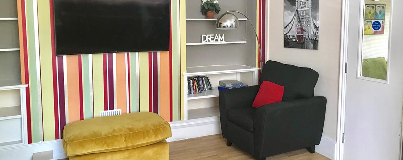 Jazzy vertical stripped wall papered wall featuring two tall alcoves with shelving and sizeable television. Large armchair and mustard footstool.