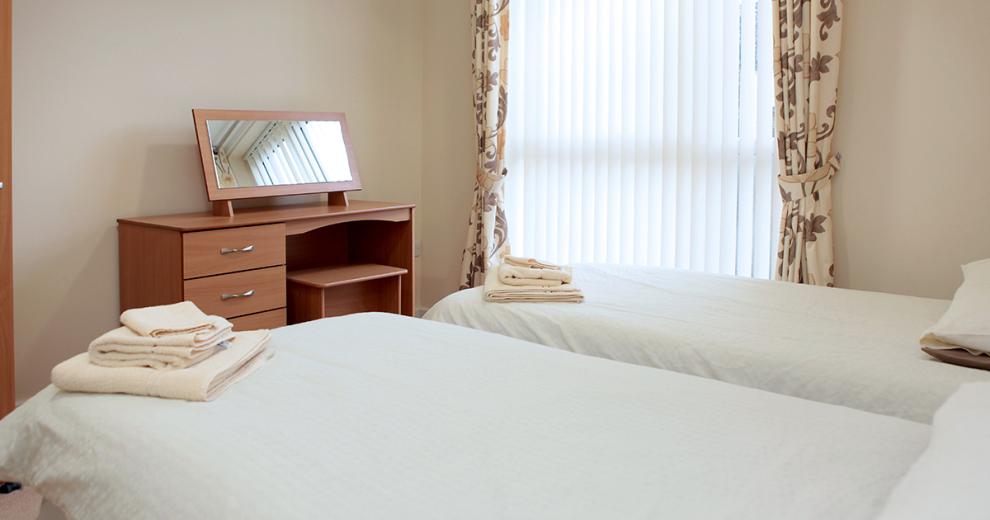 Two single beds organised centrally of a large bedroom with wooden dressing table and mirror sitting at the foot of them. A large window allowing natural light into the room is decorated with white blinds as well as green and brown leaf cream curtains.