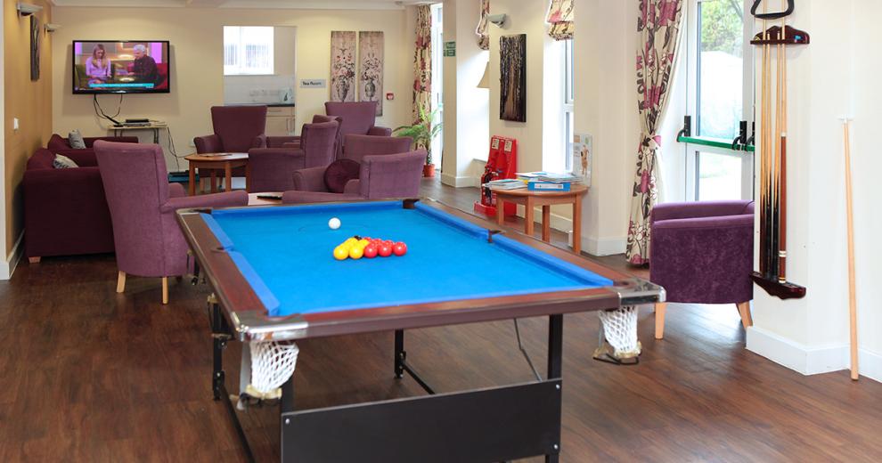 Aubergine large armchairs surround dark circular tables in front of a wall mounted television. A collapsible blue felted pool table fills the top of the room with wall mounted pool cues.