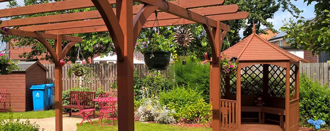 An inviting garden area with gravelled walkways leading to an open bench and dining tabled seating area, following round to a covered wooden pergola.