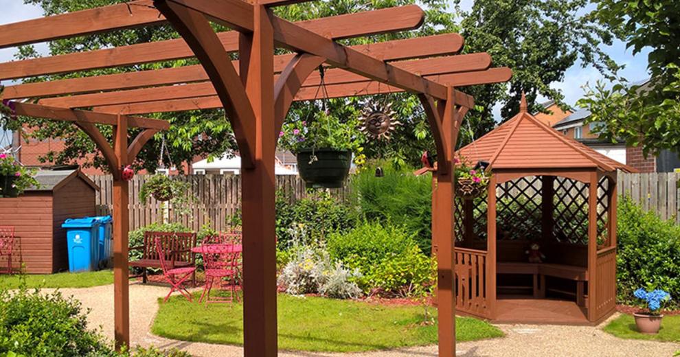 An inviting garden area with gravelled walkways leading to an open bench and dining tabled seating area, following round to a covered wooden pergola.