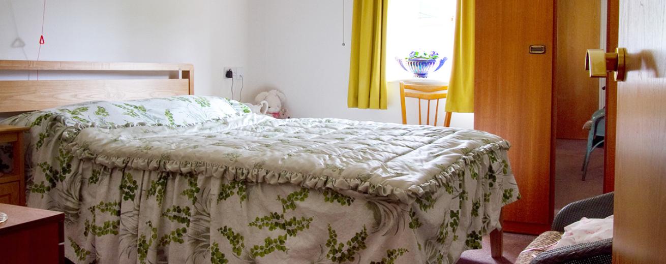 Comfortable double bedroom with minimalist wooden headboard a white and green leaf patterned bedspread, matching wooden bedroom furniture such as double wardrobe, bedside tables a chair placed under the window and a wicker chair at the foot of the bed.