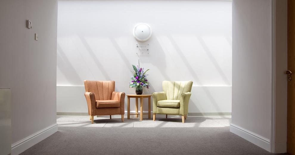 Calm and tranquil seating area, with white decor and a flooding of natural light from the skylights. Two comfortable brown and pale green leather arm chairs are placed around a small circular coffee table with floral display.
