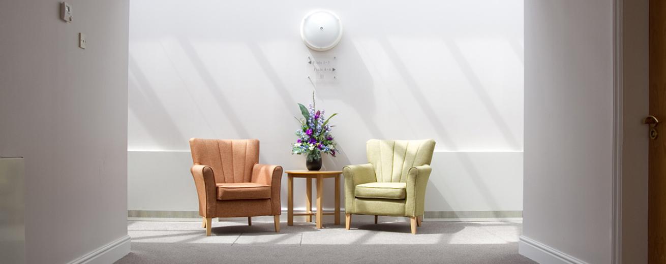 Calm and tranquil seating area, with white decor and a flooding of natural light from the skylights. Two comfortable brown and pale green leather arm chairs are placed around a small circular coffee table with floral display.