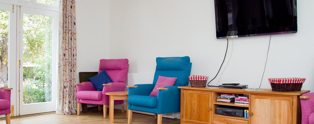 Eye catching fuchsia and blue arm chairs scattered around a neutral room with French doors leading into the garden area. The wooden flooring complements the  oak coloured wooden television unit which sits below the large wall mounted television.