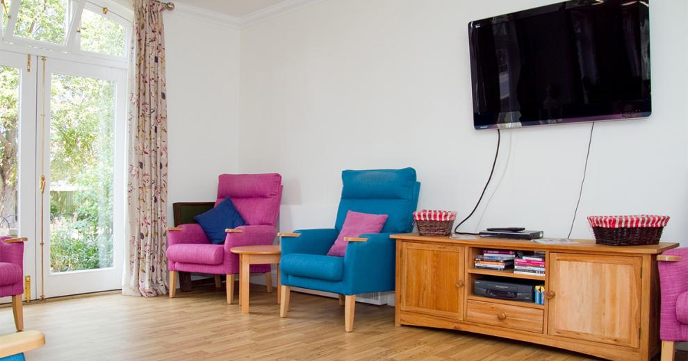 Eye catching fuchsia and blue arm chairs scattered around a neutral room with French doors leading into the garden area. The wooden flooring complements the  oak coloured wooden television unit which sits below the large wall mounted television.