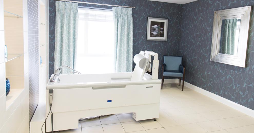 A navy wallpaper with light blue interconnecting leaf pattern, breaks up a large light and airy white tiled bathroom space, lit with natural light from a large floor to ceiling window. The room has an assisted bathing bath fitted.