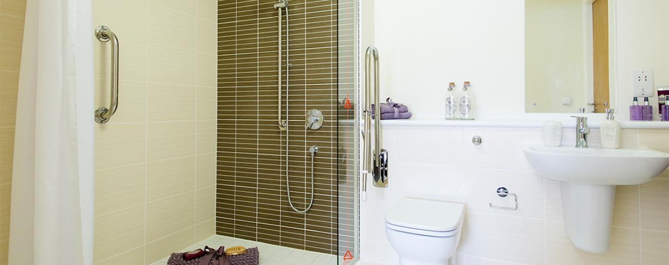 A minimalistic en-suite bathroom decorated with large white tiles and matching white paintwork, broke up by a brown tiled feature wall for the walk-in shower head to be mounted onto. The bathroom suite of toilet and sink follows the same white minimalistic design, finished off with chrome finishes and a mix of luxurious toiletries.