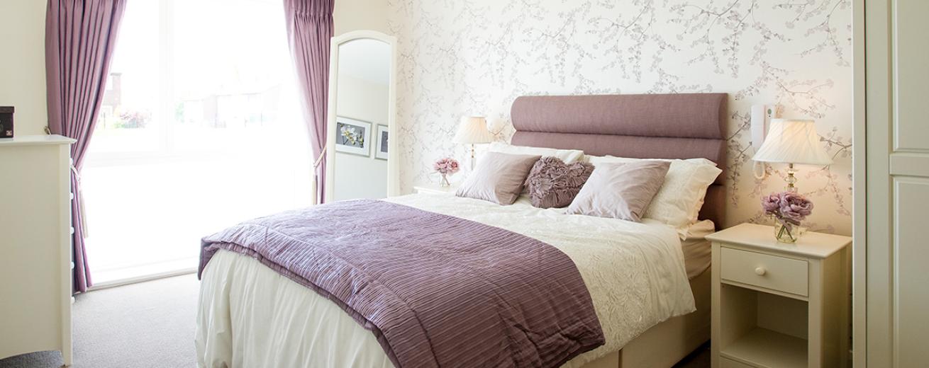 Peaceful double bedroom, tastefully decorated with delicate lilac blossom flowers on a cream background. Generous sized window allowing an abundance of light into the room. White free-standing mirror and matching white furniture set consisting of two bedside tables, a chest of draws and wardrobe add functionality with a clean design.